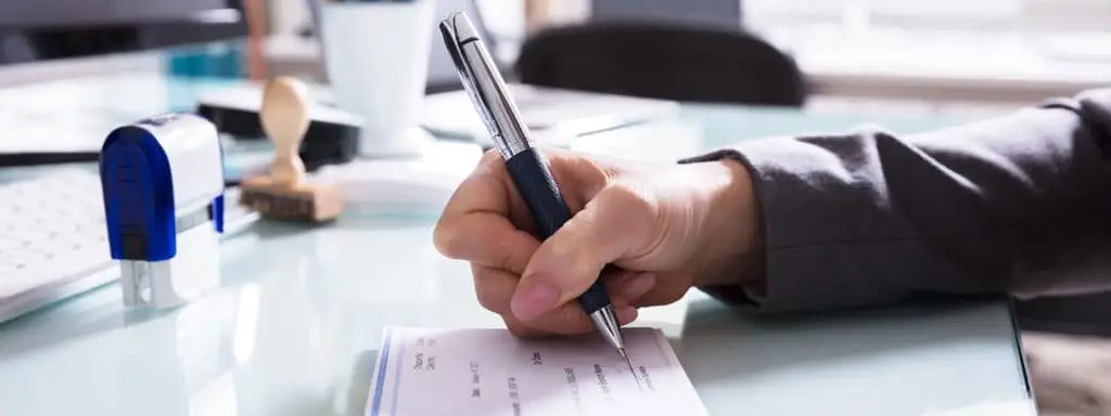 Primer plano de la mano de una persona escribiendo en un cheque con un bolígrafo en un escritorio.