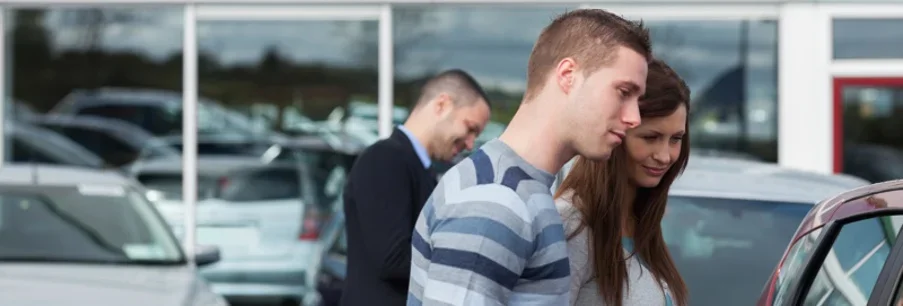 Un vendedor de coches sonriente entrega las llaves a una pareja feliz junto a un coche blanco nuevo en la sala de exposición de un concesionario.